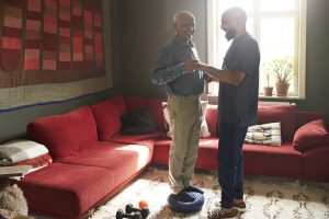 Smiling senior man taking support of male caregiver while exercising in living room